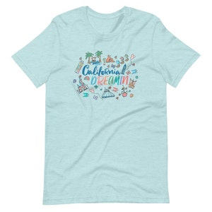 May include: Light blue t-shirt with a colorful graphic design featuring the text "California Dreamin'" and various cartoon images of attractions and landmarks.