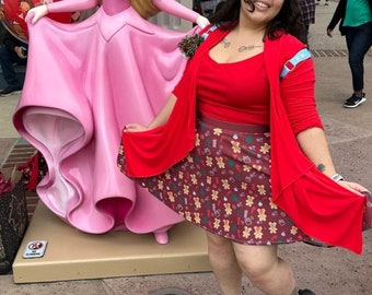 May include: A woman wearing a red cardigan over a red top and a brown skirt with a gingerbread cookie pattern. She is standing in front of a large pink statue of a princess.