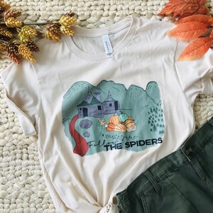 May include: A cream-colored short-sleeve t-shirt featuring a graphic print of a stone cottage, a red path, and a pile of orange and green pumpkins. The text "FOLLOW THE SPIDERS" is printed in black below the illustration, set against a sage green background.