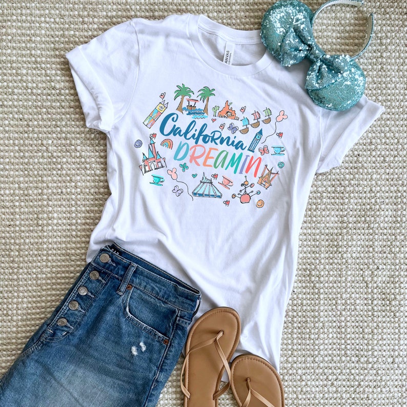 May include: White t-shirt with a colorful graphic design featuring the text "California Dreamin'" and various Disney-themed illustrations, including a castle, a train, and a Mickey Mouse head. The shirt is paired with blue jeans and brown sandals.