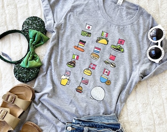 May include: A gray t-shirt with a graphic of flags and food from different countries. The flags include Japan, Germany, Italy, the Netherlands, Finland, Canada, the United Kingdom, and the United States. The food includes sushi, a pretzel, pasta, a hamburger, a cinnamon roll, poutine, fish and chips, and a hamburger.
