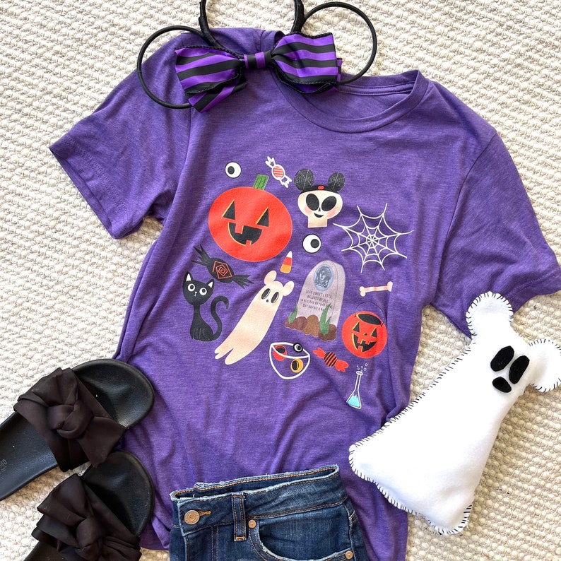 K&ouml;nnte beinhalten: Ein lila T-Shirt mit Halloween-Motiven, darunter ein K&uuml;rbis, ein Geist und ein Spinnennetz. Ein schwarz-lila gestreiftes Schleifen-Stirnband, schwarze Sandalen und ein wei&szlig;es Geisterpl&uuml;schtier sind ebenfalls zu sehen.
