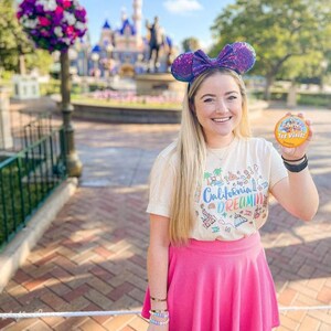 May include: A woman wearing a white t-shirt with a colorful graphic of California landmarks and the text "California Dreamin'" and a pink skirt. She is holding a round button that says "1st Visit!" and has on a sparkly purple Minnie Mouse ear headband. The background is a blurred image of a castle.