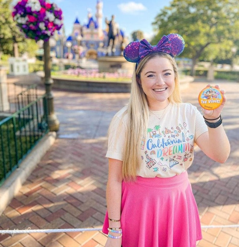 May include: A woman wearing a white t-shirt with a colorful graphic of California landmarks and the text "California Dreamin'" and a pink skirt. She is holding a round button that says "1st Visit!" and has on a sparkly purple Minnie Mouse ear headband. The background is a blurred image of a castle.