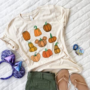 Op de afbeelding: Een crèmekleurig T-shirt met korte mouwen en een grafische print van diverse oranje pompoenen met groene steeltjes, waaronder één pompoen in de vorm van muizenoortjes. Het shirt wordt getoond met paarse paillettenoren, een olijfgroene short en sandalen.