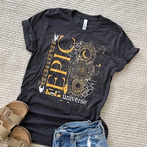 Może przedstawiać: Szary t-shirt z nadrukiem w kolorze złotym przedstawiającym słowo "EPIC" i tekst "universe" poniżej. Nadruk zawiera półksiężyc, gwiazdy i koło z wzorem.