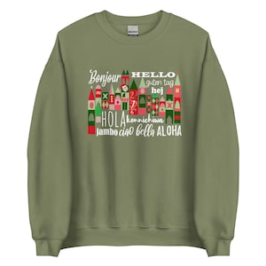 Peut inclure: Un sweat-shirt vert olive avec un motif graphique comportant le mot "Bonjour" et d'autres salutations dans différentes langues. Le motif comprend des bâtiments colorés et stylisés et le mot "HELLO" en blanc.