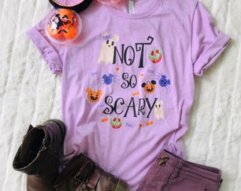 Może przedstawiać: Liliowa koszulka z napisem "NOT SO SCARY" i grafiką o tematyce Halloween. Koszulka jest zestawiona z brązowymi butami, fioletowymi spodniami i akcesoriami na Halloween. Rękawy koszulki są podwinięte.