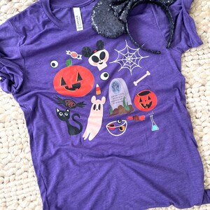 K&ouml;nnte beinhalten: Ein lila T-Shirt mit einem Halloween-Motiv mit K&uuml;rbis, Geist und anderen gruseligen Elementen. Ein schwarzes paillettenbesetztes Minnie Mouse Ohren-Stirnband liegt auf dem Shirt. Das Shirt besteht aus einem weichen Material.