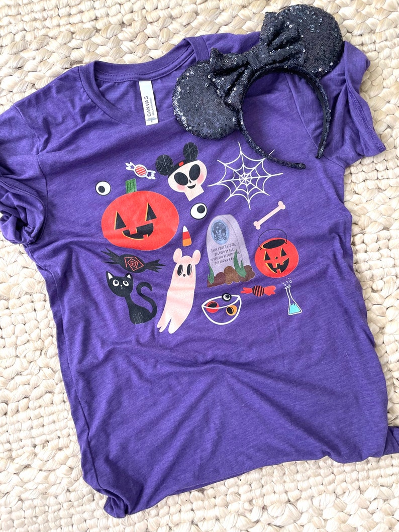 K&ouml;nnte beinhalten: Ein lila T-Shirt mit einem Halloween-Motiv mit K&uuml;rbis, Geist und anderen gruseligen Elementen. Ein schwarzes paillettenbesetztes Minnie Mouse Ohren-Stirnband liegt auf dem Shirt. Das Shirt besteht aus einem weichen Material.