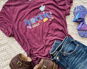 May include: A maroon t-shirt with the word "Foodie" in blue, featuring food-related graphics. The shirt is paired with blue denim jeans, brown ankle boots, and a sequined purple headband with a bow. The items are arranged on a white rug.