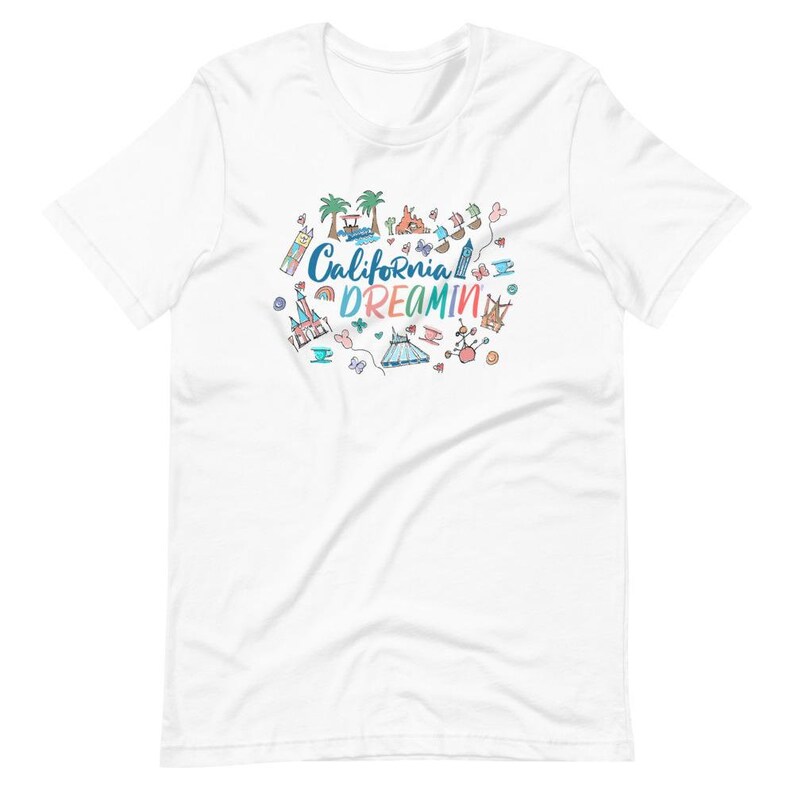 May include: White t-shirt with a colorful graphic design featuring the text "California Dreamin'" and various cartoon images of amusement park attractions, including a castle, a roller coaster, and a carousel.
