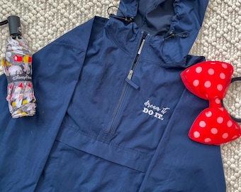 May include: A navy blue hooded windbreaker with a half-zip and the text "dream it DO IT." An umbrella with a Disney Parks design and a red polka dot bow are also in the image.