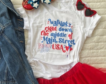 Può includere: Maglietta bianca con una grafica blu e rossa che dice "Walking right down the middle of Main Street USA". La grafica include fuochi d'artificio e un palloncino di Topolino. La maglietta è abbinata a una gonna di tulle rossa e una giacca di jeans.