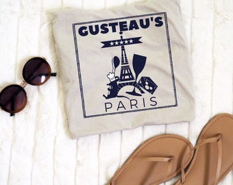 May include: A beige t-shirt with a navy blue graphic that reads "GUSTEAU'S PARIS" with an illustration of the Eiffel Tower, a chef, and cheese. The image also includes brown sunglasses and tan flip-flops.