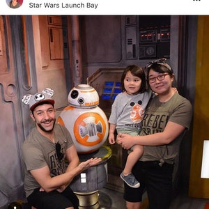 May include: A family poses for a photo with a Star Wars BB-8 droid at Star Wars Launch Bay. The family includes a man wearing a black and white Mickey Mouse ears hat, a woman wearing a grey shirt with the word "Rebel" on it, and a young child wearing a grey shirt with a BB-8 graphic.