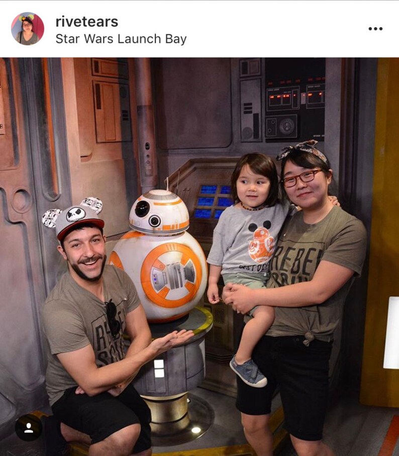 May include: A family poses for a photo with a Star Wars BB-8 droid at Star Wars Launch Bay. The family includes a man wearing a black and white Mickey Mouse ears hat, a woman wearing a grey shirt with the word "Rebel" on it, and a young child wearing a grey shirt with a BB-8 graphic.