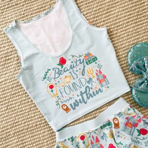 May include: A light blue tank top with a white background and the text "Beauty is found within" in a whimsical font. The tank top is decorated with illustrations of roses, a teapot, a clock, a castle, and books.