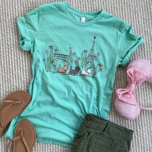 Puede incluir: Una camiseta verde claro con un gráfico de tres gatitos caminando frente a la Torre Eiffel. Los gatitos llevan collares y caminan por un camino con flores y un puente. La camiseta tiene una manga enrollada.
