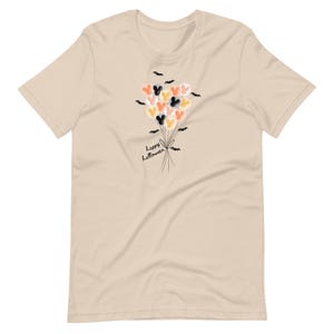 May include: Beige t-shirt with a design of Mickey Mouse-shaped balloons in orange, black, and yellow. The balloons are tied with a ribbon and surrounded by bat silhouettes. The words "Happy Halloween" are written below.