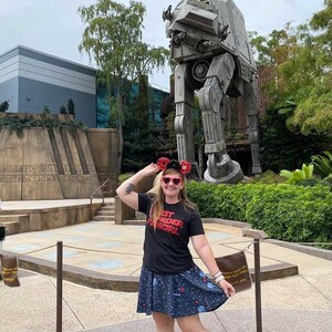May include: A woman wearing a black t-shirt with the text "Best Order Fangirl" in red letters, a blue skirt with white stars and a red bow, and red Mickey Mouse ears, poses in front of a large, grey, mechanical walker.