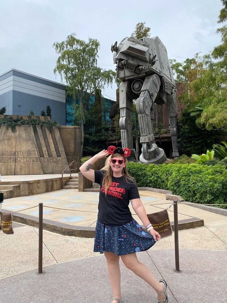 May include: A woman wearing a black t-shirt with the text "Best Order Fangirl" in red letters, a blue skirt with white stars and a red bow, and red Mickey Mouse ears, poses in front of a large, grey, mechanical walker.