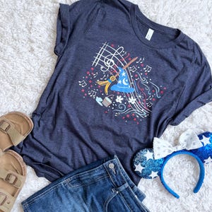 May include: Navy blue t-shirt with a wizard-themed graphic, musical notes, and stars. The shirt is styled with blue denim shorts, sandals, and sequined mouse ears. A casual outfit, perfect for a theme park.