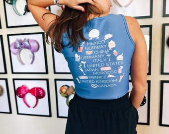 May include: A light blue crop top with white graphic icons and text listing countries like Mexico, Norway, and China. The top is paired with black shorts. Framed decorative headbands are in the background.