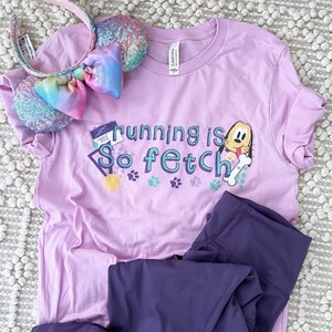 May include: A lilac t-shirt with the text "Running is So Fetch" and a cartoon dog graphic, paired with purple leggings and a sequined headband with a rainbow bow. The shirt has paw prints and a gold medal graphic.