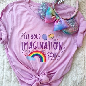 May include: A pink t-shirt with the text "Let your imagination soar" printed on it. The text is in purple and is surrounded by a rainbow, a cloud, a music note, and a paint palette.