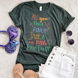 May include: A green t-shirt with the text "All you need is faith, trust, & a little pixie dust" in colorful lettering. The shirt is laid out on a white surface with a pair of sandals, a pair of white denim shorts, and a pair of Minnie Mouse ears.