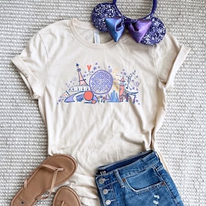 Puede incluir: Camiseta beige con un gráfico colorido de varios monumentos y atracciones. El diseño incluye una gran esfera morada, una torre y otros elementos arquitectónicos. La camiseta se combina con pantalones cortos de mezclilla, sandalias y una diadema con lentejuelas y un lazo.