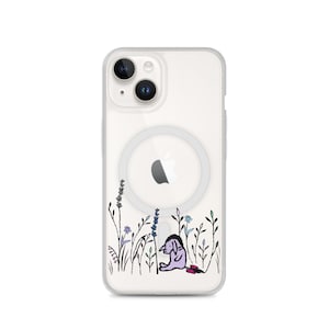 May include: Clear phone case with a cartoon illustration of a purple donkey sitting in a field of flowers. The case has a MagSafe ring and an Apple logo. The flowers are in shades of blue, purple, and green.