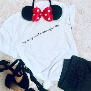 May include: A white t-shirt with the text "my oh my what a wonderful day" in black script. A pair of black mouse ears with a red bow with white polka dots sits on top of the shirt. Black pants and shoes are also in the image.