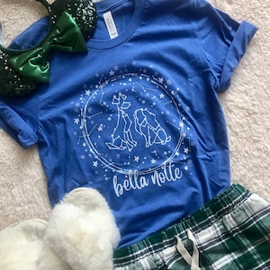 Puede incluir: Una camiseta azul con un dise&ntilde;o blanco de dos perros en un c&iacute;rculo de estrellas. El texto "bella notte" est&aacute; debajo del dise&ntilde;o.