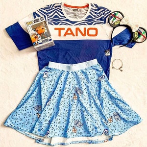 Puede incluir: Una camiseta a rayas azul y blanca con la palabra "TANO" en letras naranjas. La camiseta se combina con una falda azul con un estampado de estrellas blancas y azules y un peque&ntilde;o personaje de la franquicia Star Wars. La falda es de estilo patinadora con un forro blanco.