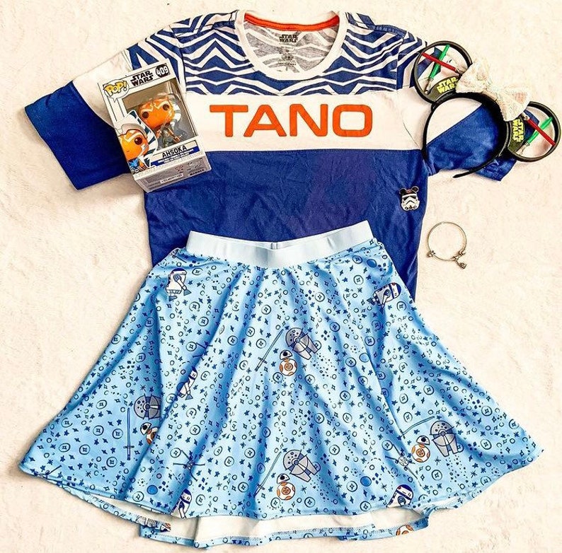 Puede incluir: Una camiseta a rayas azul y blanca con la palabra "TANO" en letras naranjas. La camiseta se combina con una falda azul con un estampado de estrellas blancas y azules y un peque&ntilde;o personaje de la franquicia Star Wars. La falda es de estilo patinadora con un forro blanco.