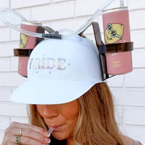 May include: White baseball cap with the word "BRIDE" in silver glitter letters. The cap has two clear plastic tubes attached to it, each connected to a pink can with a lemon graphic.