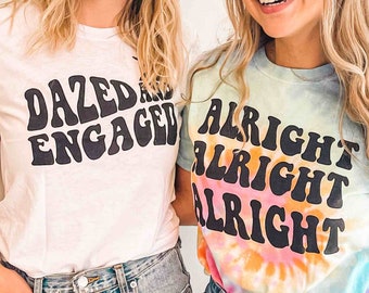 May include: Two women wearing white and tie-dye t-shirts with text. The white shirt says "Dazed and Engaged" in a retro font. The tie-dye shirt says "Alright Alright Alright" in a retro font.