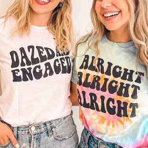 May include: Two women wearing white and tie-dye t-shirts with text. The white shirt says "Dazed and Engaged" in a retro font. The tie-dye shirt says "Alright Alright Alright" in a retro font.