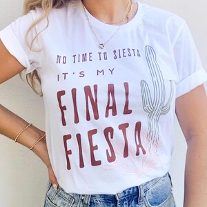 May include: White t-shirt with rolled-up sleeves featuring the text "No Time to Siesta, It's My Final Fiesta" in burgundy. A cactus and floral design are printed on the shirt. The model is wearing blue jeans and gold jewelry.