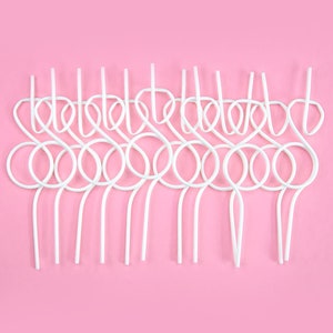 May include: A collection of white, looped straws arranged in a row against a solid pink background. The straws have a unique design with a series of circles and vertical lines, creating a decorative pattern. The image is a top-down view.