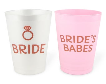 May include: Two plastic cups, one white and one pink. The white cup has a ring graphic and the word "BRIDE" in copper-colored text. The pink cup has the words "BRIDE'S BABES" in the same copper color. Perfect for a bridal shower.