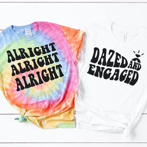 May include: Two tie-dye t-shirts with a retro font design. One shirt is a rainbow tie-dye with the text "Alright Alright Alright". The other shirt is white with the text "Dazed and Engaged".