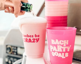 May include: Pink plastic cups with text that reads "Baches be CRAZY" and "BACH PARTY GOALS". A bottle of champagne is pouring into one of the cups, creating foam. A stack of pink cups is in the background.