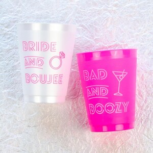 May include: Two plastic cups with pink lettering. One cup is white with the text "Bride and Boujee" and a diamond ring icon. The other cup is pink with the text "Bad and Boozy" and a martini glass icon.