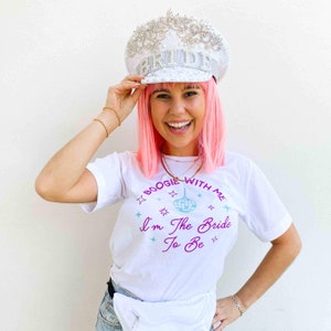 May include: A white t-shirt with the text "Boogie With Me I'm The Bride To Be" in purple and blue. The image also features a white, embellished "BRIDE" hat with silver accents.
