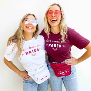 Op de afbeelding: Twee vrouwen dragen hartvormige zonnebrillen en t-shirts met de tekst "HERE COMES THE BRIDE" en "HERE COMES THE PARTY". Ze dragen ook heuptasjes die passen bij het thema van het shirt. De shirts zijn wit en bordeauxrood.