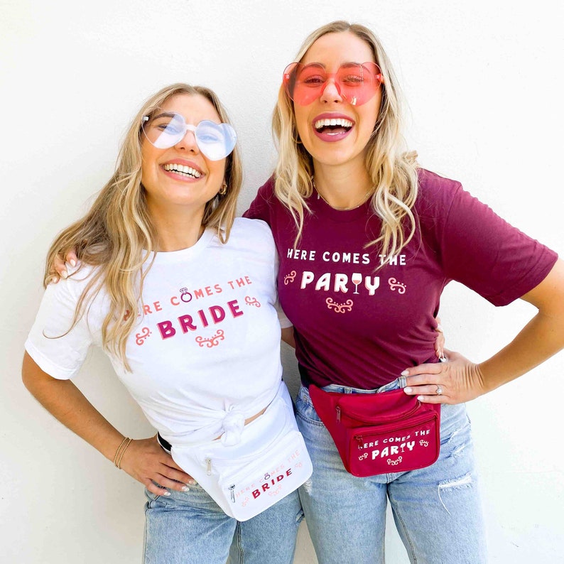 Op de afbeelding: Twee vrouwen dragen hartvormige zonnebrillen en t-shirts met de tekst "HERE COMES THE BRIDE" en "HERE COMES THE PARTY". Ze dragen ook heuptasjes die passen bij het thema van het shirt. De shirts zijn wit en bordeauxrood.