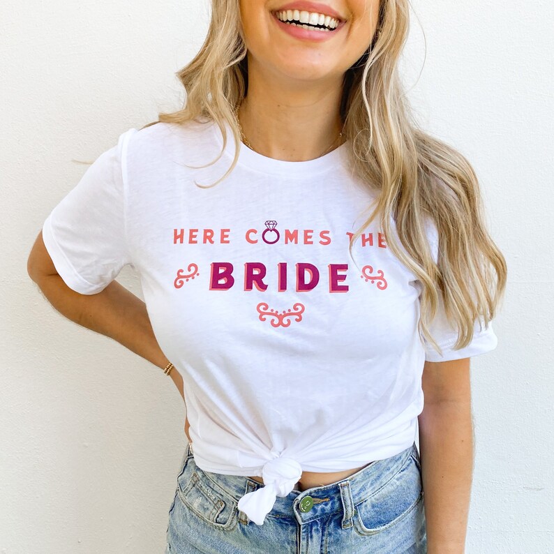 Op de afbeelding: Wit T-shirt met de tekst "HERE COMES THE BRIDE" in roze en magenta. Het ontwerp bevat een diamantring-afbeelding en decoratieve krullen. Het shirt is in de taille geknoopt.
